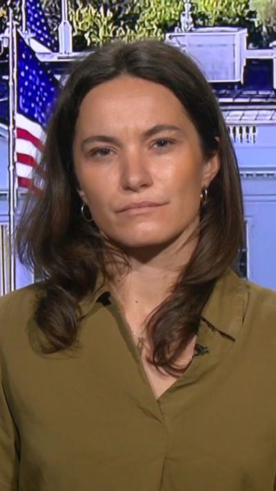 Ione Wells looks into the camera in front of a digital screen showing the white house, she wears a green top.