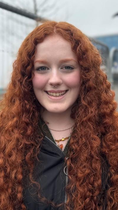 A young woman with long, wavy red hair looks into the camera. She is smiling and has a silver hooped nose ring.