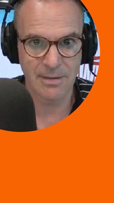 Martin Lewis wears black shirt with white stripes and glasses. He sits behind a microphone