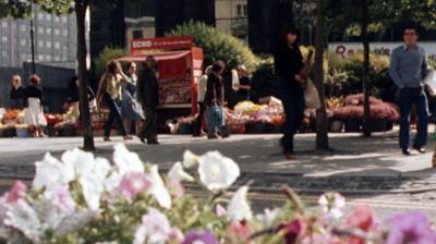 Low angle shot of people walking along the pavement in Liverpool past a bed of flowers and a Liverpool Echo branded magazine stand. 