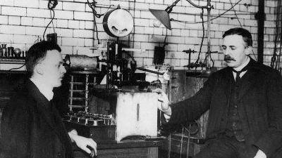 Ernest Rutherford (right) and Hans Geiger sit either side of a table of lab equipment in a black-and-white photo of a physics laboratory in Manchester in 1913.