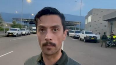 A BBC reporter looks into the camera at dusk in front of a border checkpoint, two men are seen in the background..