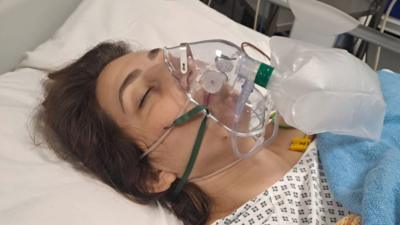 Brunette woman lying down in hospital bed with oxygen mask over her face and eyes closed.