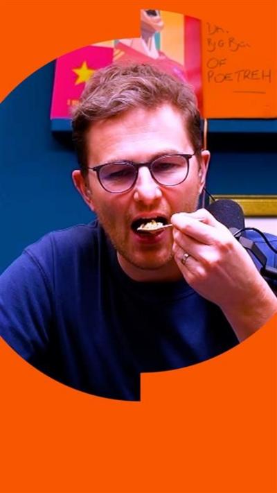 A man with a blue t-shirt and glasses, holding a spoon going into his mouth, in a podcast studio.