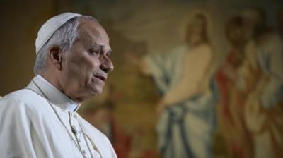 Pope Leo in the Vatican, dressed in papal robes. In front of a big TV showing Americans.