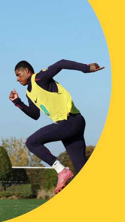 A man, Marcus Rashford, wears a black tracksuit with a yellow high vis vest and football boots. He is running.