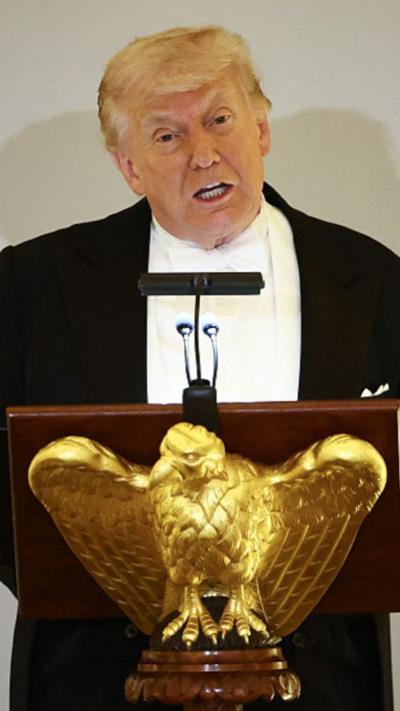 President Donald Trump, dressed in a black tuxedo with a white shirt and tie, addresses a room behind a podium with a golden eagle.