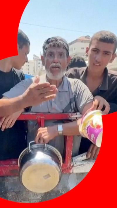 Tayseer Naeem, an older man with a grey beard, holds a saucepan amidst a line of people behind a metal fence at a soup kitchen in Gaza.