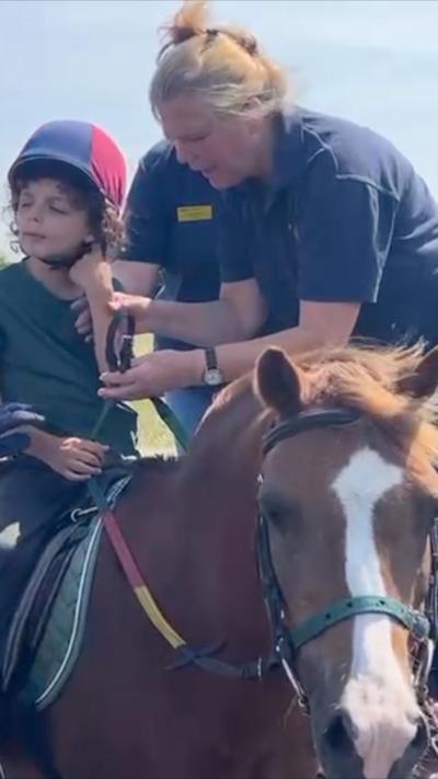 Young boy in a green top sits on the back of a pony, a woman with blond hair tied up wearing a blue polo shirt stands to his side