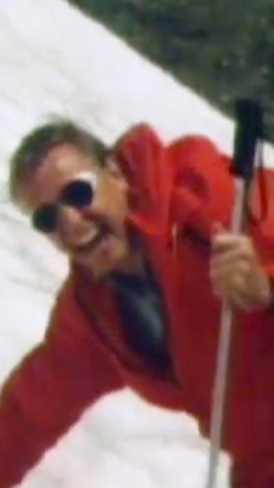 Attenborough, in a bright red coat and sunglasses, laughs as he slides down a steep snowy slope, clutching a walking pole