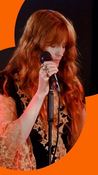 Florence Welch stands behind a mic with her eyes closed. She wears a black and gold top 