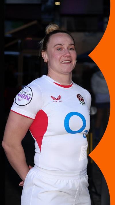 A woman in a white England rugby shirt stands next to a trophy.