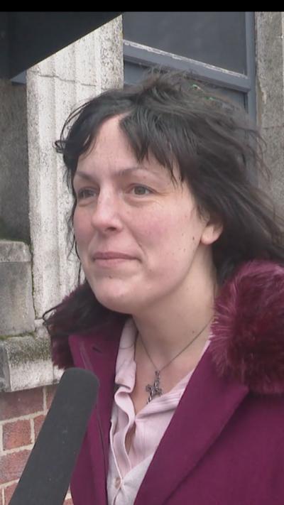 A woman with long dark hair speaks into a microphone. She is wearing a pink blouse and a maroon coat with a furry hood.