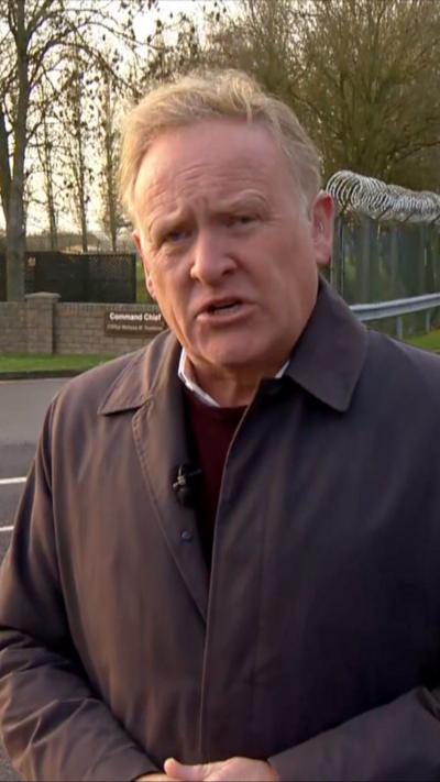 BBC journalist John Maguire stands outside RAF Fairfood. He is wearing a light jacket which has a microphone attached. Barbed wire can be seen around the entrance to the air base.