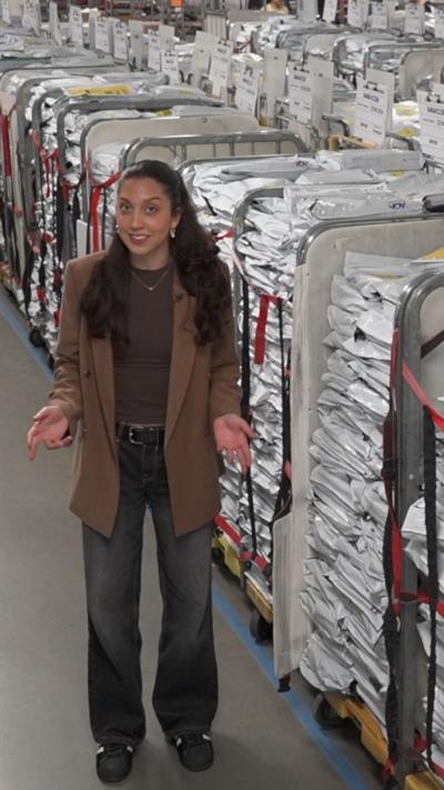 A woman in a brown coat and jeans stands next to a number of large trolleys containing exam papers.