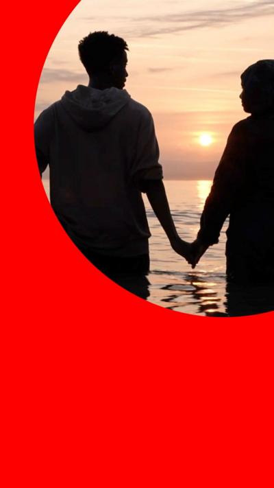 Couple stand in the sea, holding hands, with the Sun in the background 