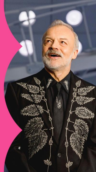Graham Norton stands on the Doctor Who set; he wears a black blazer, shirt and tie with embroidered leaves