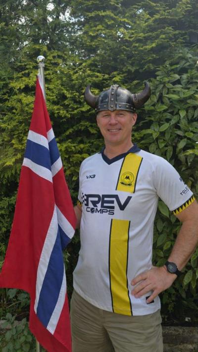 Man in a white and yellow Torquay United football top wears a Viking helmet and holds a red and blue and white Norweigan flag in front of a green hedge 