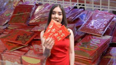 Sabrina Lee wears a red dress and holds red money packets with Chinese writing on them. A picture of many of these money packets is seen behind her.