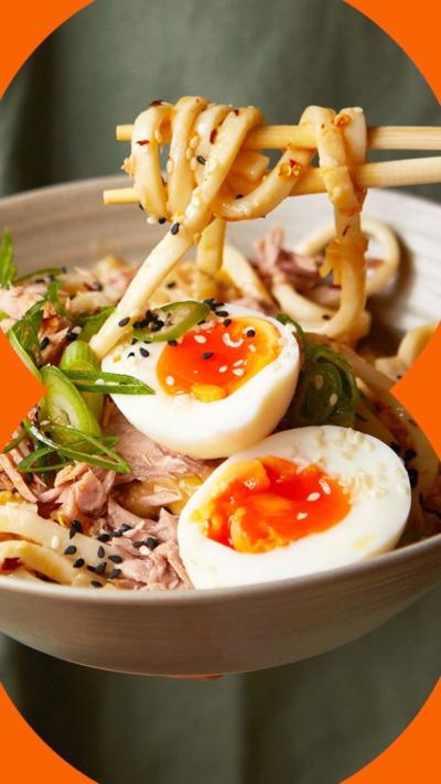 A bowl of udon noodles with sliced boiled egg and tuna on the top, with someone holding chopsticks holding some of the noodles.