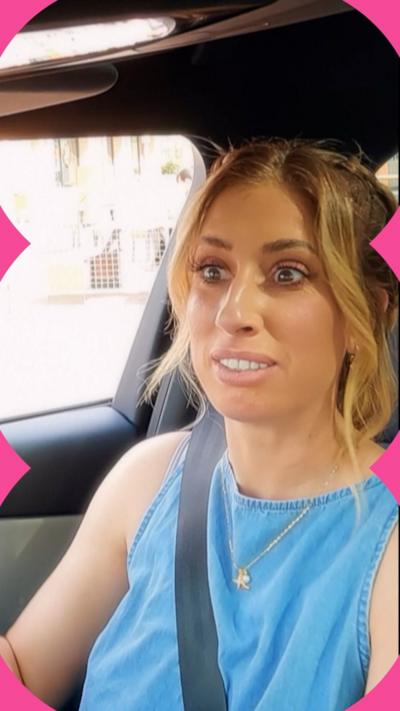 A woman with dark blonde hair wearing a blue vest top sits in the front seat of a car.