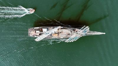 A tall ship photographed from above. The huge sails are shown and it is moving forward through the water which has a green hue to it. There is a smaller orange speedboat behind it.