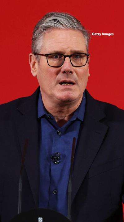 A man with grey hair and glasses, wearing a blue shirt, standing infront of a bright red background