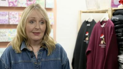 Blonde woman smiling, wearing a denim shirt and standing in front of a wall of cards and jumpers with designs on them.