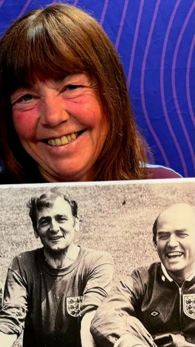 A woman with long brown hair holding up a large black and white photograph of two men in England football tracksuits