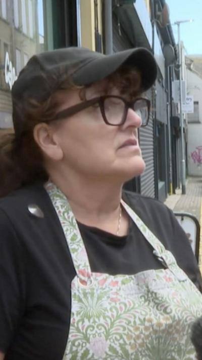 A woman with brown hair tied back in a low pony tail. She is wearing a black baseball cap and glasses with a dark frame. Her black t-shirt is covered by a flowery apron. 