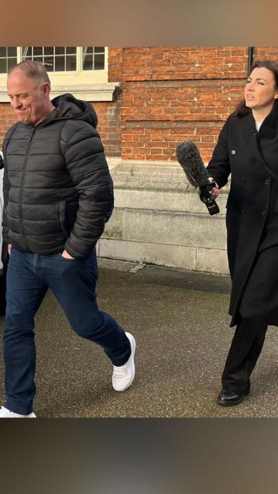 Raymond McDonald, a man in his fifties, is walking away from Philippa Goymer who is a BBC journalist. She is dressed in black and has long dark hair and is carrying a large microphone. He has a black jacket, blue jeans and has his hands in his pockets. Behind them both is a brick built building and a white car is parked to the side.
