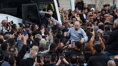 Man in prison uniform being carried in the air by crowd after leaving bus. Many people are aiming phone cameras at him