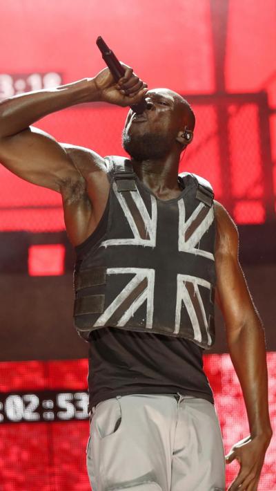 Stormzy, singing into a microphone wearing a Banksy designed bulletproof vest with a Union Jack, performs at Glastonbury 2019 
