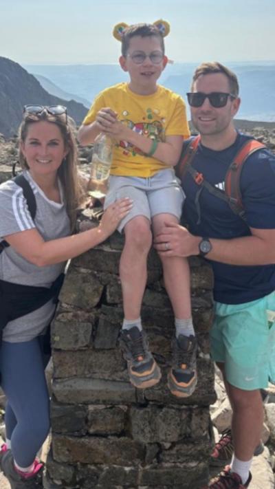 A boy in a yellow t-shirt sat on a block of stones with her mum and dad. His mum has long brown hair and is wearing a grey top and blue leggings. His dad is wearing a blue t-shirt and green shorts and sunglasses