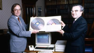 Image of two suited men holding two large laser discs above an 80s computer