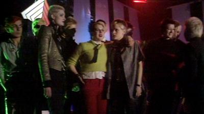 A group of people stand in a dark studio wearing punk clothing. 