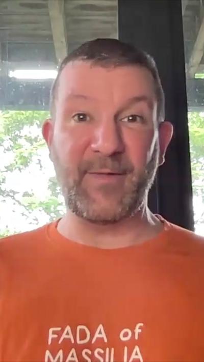 Unshaven man with short dark hair wearing a orange top with writing in the front