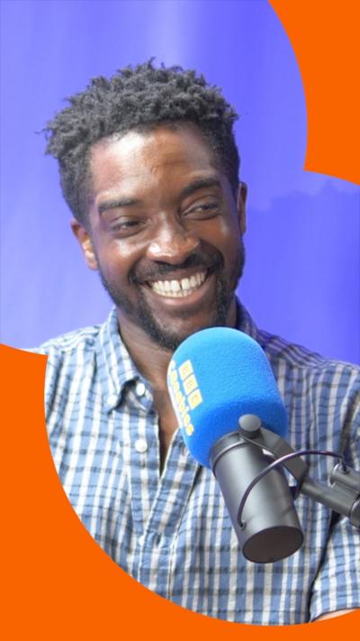 A man with short, dark hair smiles as he talks into a microphone