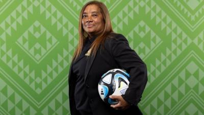 Desiree Ellis poses for official photo in suit and football tucked under one arm