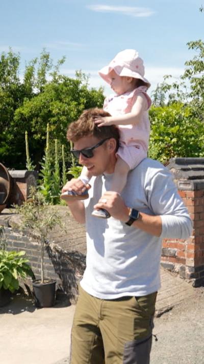 Dad walking along with daughter on his shoulders. The baby is wearing a pink outfit and pink hat. The man is wearing a blue shirt, cargo trousers and sunglasses.