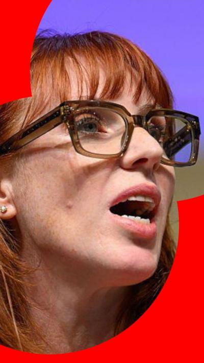 A woman with long, ginger hair and a fringe talks to someone off-camera. She wears thick-rimmed glasses