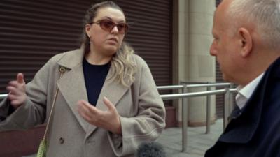 A woman in a grey coat and sunglasses talking to Steve Rosenberg
