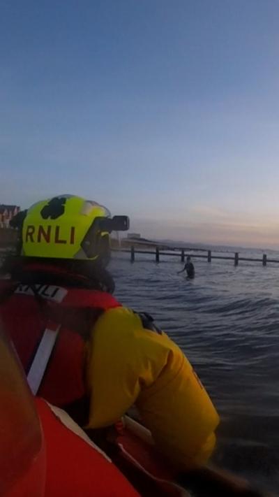 The back of an RNLI crew member in a bright yellow helmet looking out to shallow sea water where a paddler is waist deep in water. You can see a town promenade in the background and low evening light.