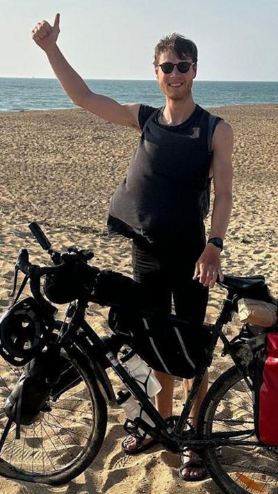 Man standing on the beach next to his bike with his arm raised giving a thumbs up