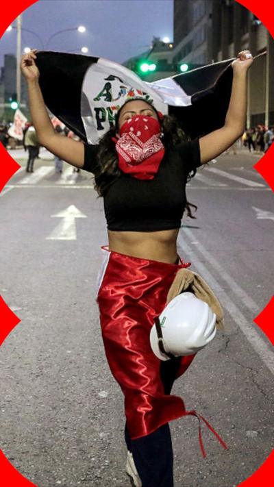 Person wearing bandana over face and red flag around her waist walking in the street and holding a black and white flag in her hands