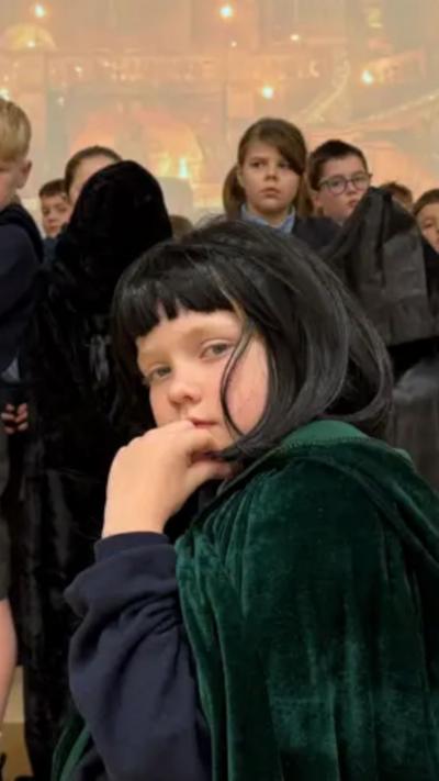A schoolboy in a black wig and green cloak with his hand on his chin in a thinking position
