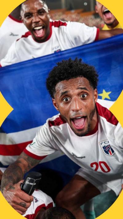 
Cape Verde footballers celebrate a victory, with captain Ryan Mendes, wearing a white shirt with red trim, crouching down and extending his arms in front of a national flag and pulling a wide-eyed face of celebration with his mouth wide open.