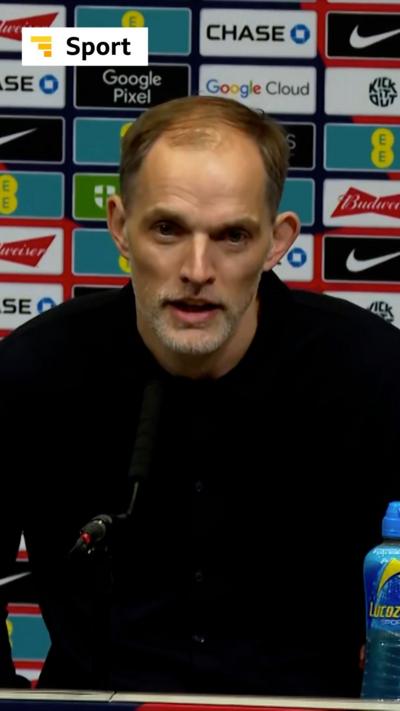 A man, Thomas Tuchel, wears a dark coloured jumper and talks into a microphone