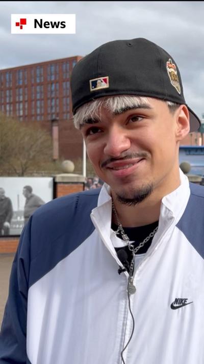 A man smiles, wearing a black baseball cap and a  white and blue sports jacket 