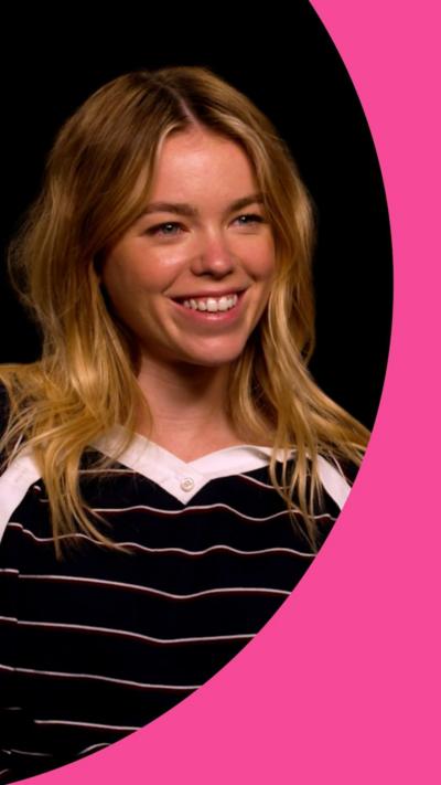 Milly Alcock has shoulder-length blonde hair. She is wearing a black top with thin horizontal white stripes.